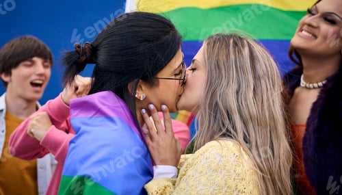 Preview: Young Caucasian lesbian couple kissing lovingly with rainbow flags celebrating with LGBT