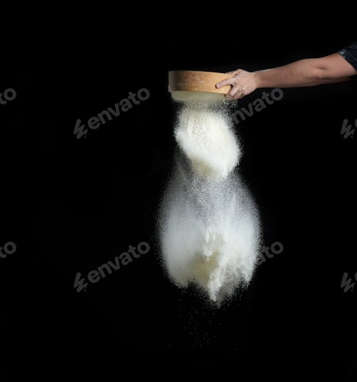 Preview: Female hand sifts white wheat flour through a round wooden sieve on a black background