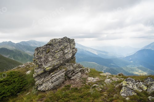 Preview: Rock on a hill covered in greenery with rocky mountains under a cloudy sky on the blurry background