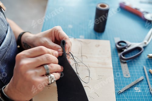 Preview: Hands of leatherworker with piece of black leather sewing new item