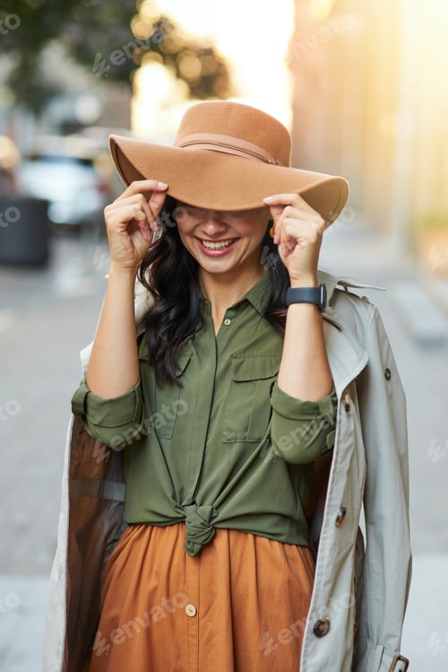 Preview: Playful beauty. Young happy caucasian woman adjusting hat and smiling while standing on the city