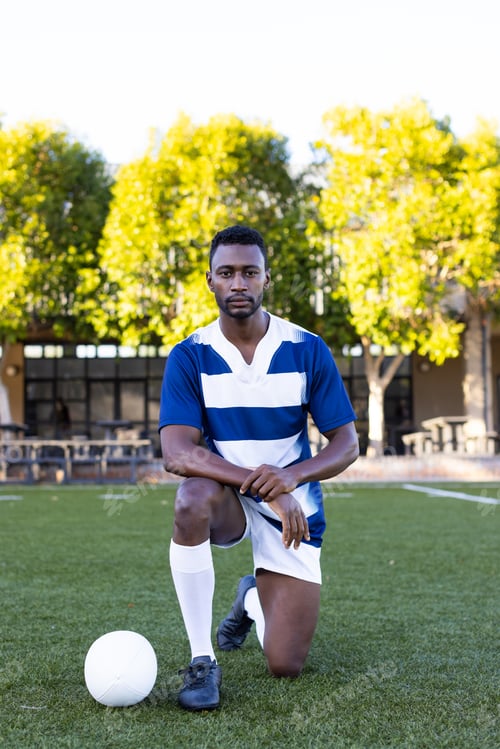 Preview: African American young male athlete kneeling next to a soccer ball on field outdoors, copy space