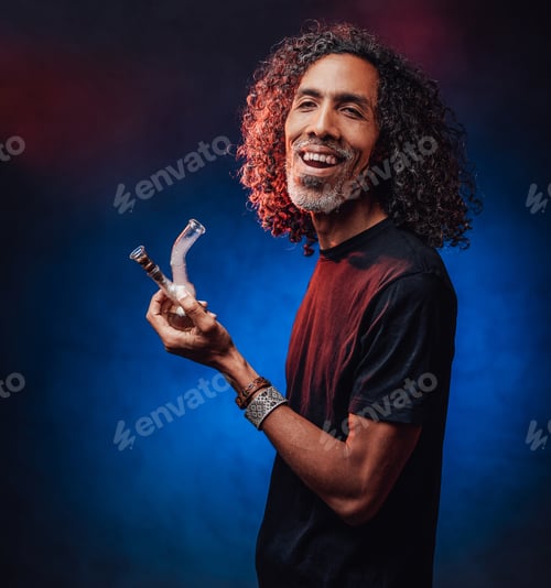 Preview: Middle aged hispanic male smoking from bong on a dark background illuminated blue light