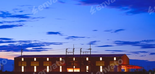 Preview: Silhouette industrial building with illuminated electric lighting inside against blue hour dusk sky