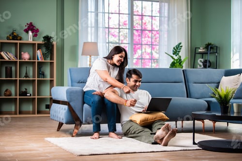 Preview: Young and happy Indian couple sitting on sofa at home, using a laptop for shopping or video call