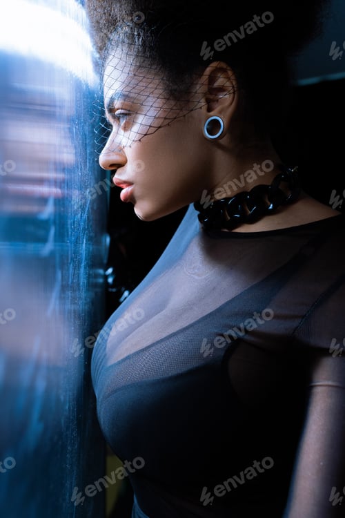 Preview: young african american woman in sexy outfit and black veil looking through glass with blue lighting