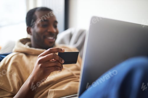Preview: Young Man At Home Lying On Sofa With Laptop Using Credit Card To Shop Online