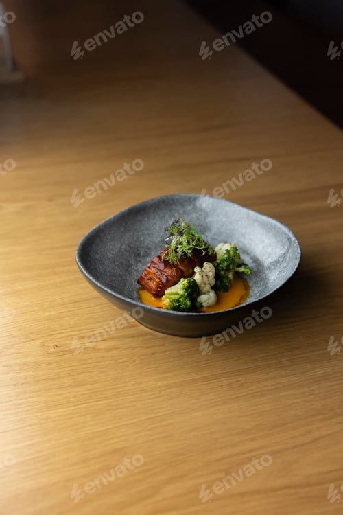 Preview: grey bowl filled with a delicious dish of roasted pork belly with cauliflower, broccoli, and a tangy