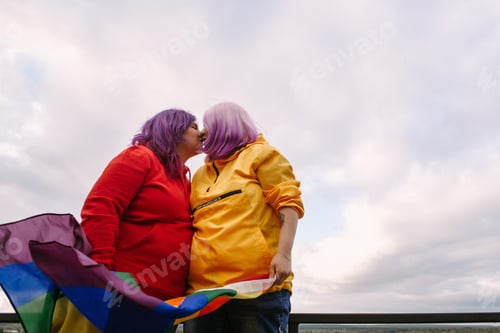 Preview: happy young women lesbian holding lgbtq pride flag and kissing