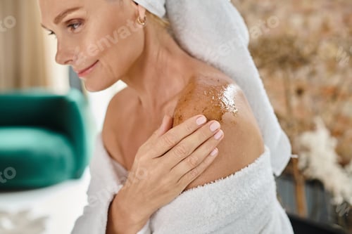 Preview: beautiful middle aged woman with white towel on head and bathrobe using body scrub on shoulder