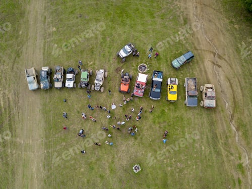 Preview: Aerial shot of lots of people near off-road vehicles in a grass field near trees