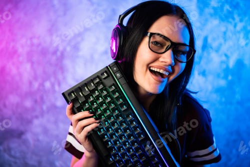Preview: Nerd geek young adult women holding gaming keyboard over colorful pink and blue neon lit wall