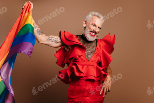 Preview: Happy mature gay man in beautiful red dress waving rainbow flag against brown background