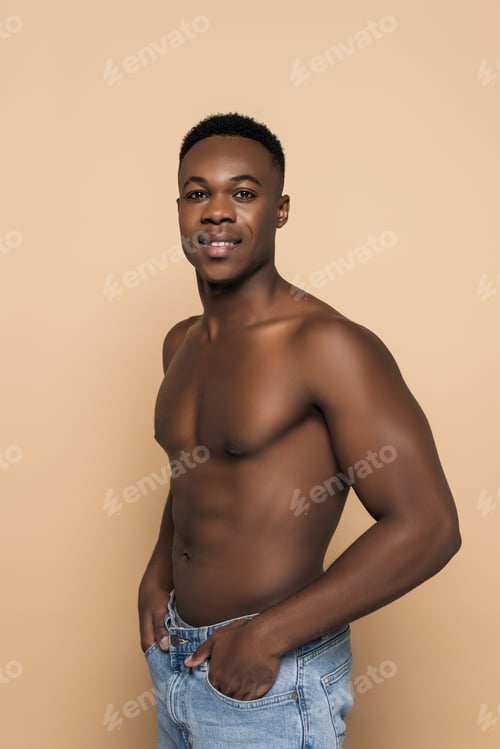 Preview: shirtless african american man smiling while standing with hands in pockets isolated on beige