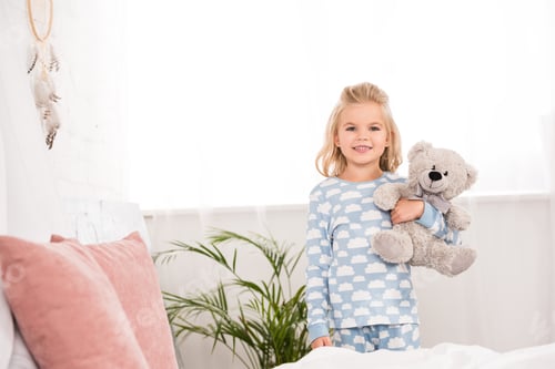 Preview: smiling child in pajamas standing with teddy bear in bedroom