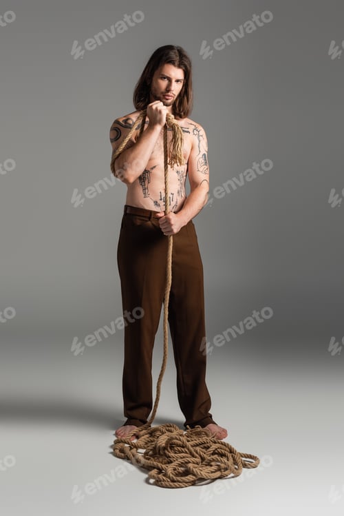 Preview: Full length of tattooed man in pants holding rope on grey background
