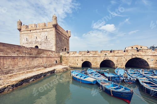 Preview: Blue boats floating by an ancient fortress under a clear sky.