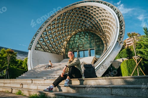 Preview: Purposeful young man sitting on stairs against modern building on sunny day dreaming about future,
