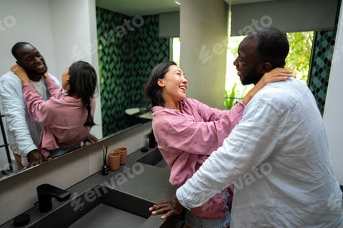 Visualização: Jovem casal diversificado sorrindo alegremente juntos em sua luminosa sala de estar, vestindo roupas casuais
