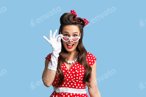 Preview: Emotional pin up woman in retro fashion dress touching sunglasses, looking at camera in excitement