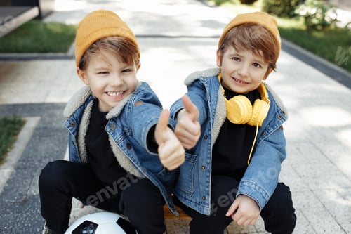 Preview: Two happy twin boys sitting on the skateboard or pennyboard showing thumbs up in the street.