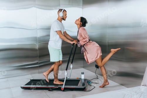 Preview: woman in robe kisses a man on treadmill. The man wearing headphones and exercise clothes, highlighti