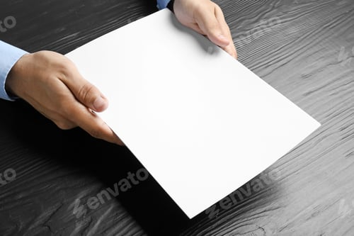 Preview: Man holding blank paper sheets for brochure at black wooden table. Mock up