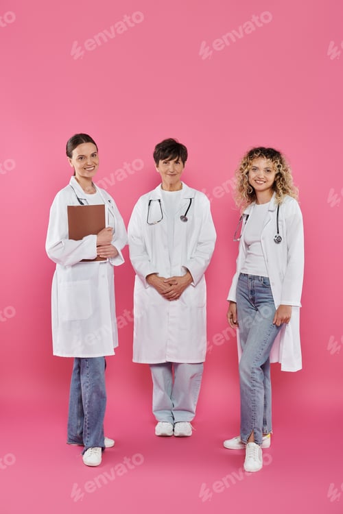 Preview: smiling female doctors holding paper folder and looking at camera on pink, breast cancer concept