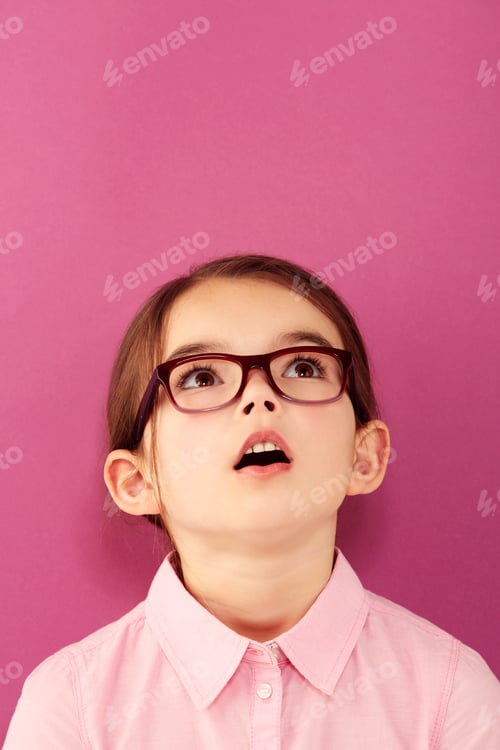 Preview: Childlike wonder. A little girl wearing spectacles looking up against a pink background.