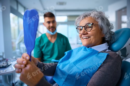 Preview: Satisfied senior woman using mirror while checking her teeth after procedure at dentist's office.