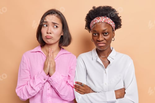 Preview: Upset dark haired woman keeps palms pressed together in pray looks with pleading expression dressed