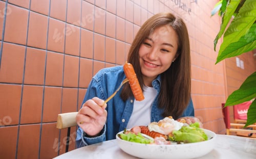 Preview: Portrait image of a young woman showing a sausage while eating salad