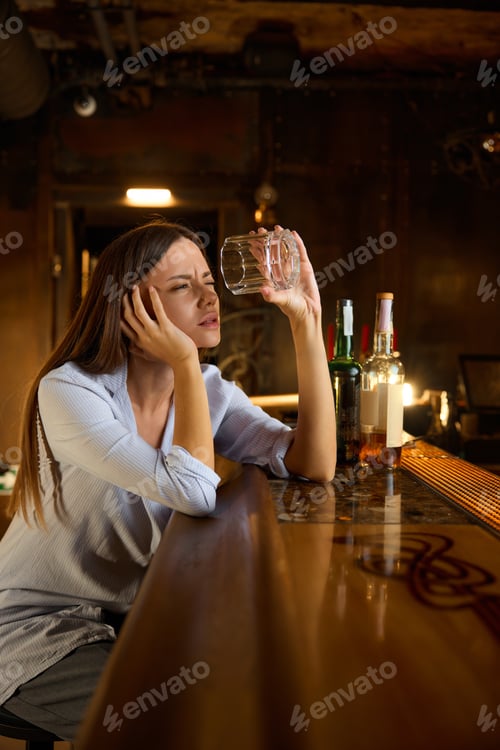 Preview: Drunk young woman looking into empty glass sitting at bar counter