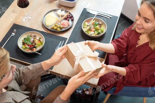 Preview: Hands of young man passing giftbox with white silk ribbon to his girlfiend