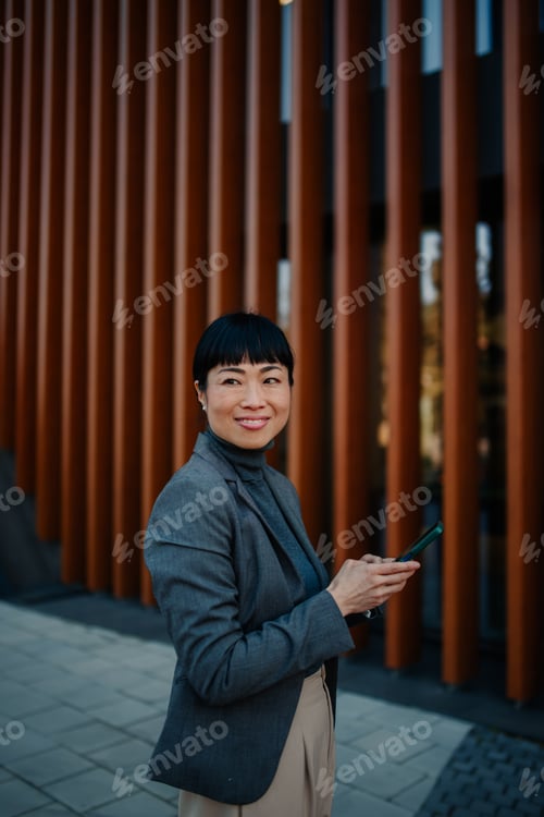 Preview: Smiling businesswoman using smartphone in front of modern office building