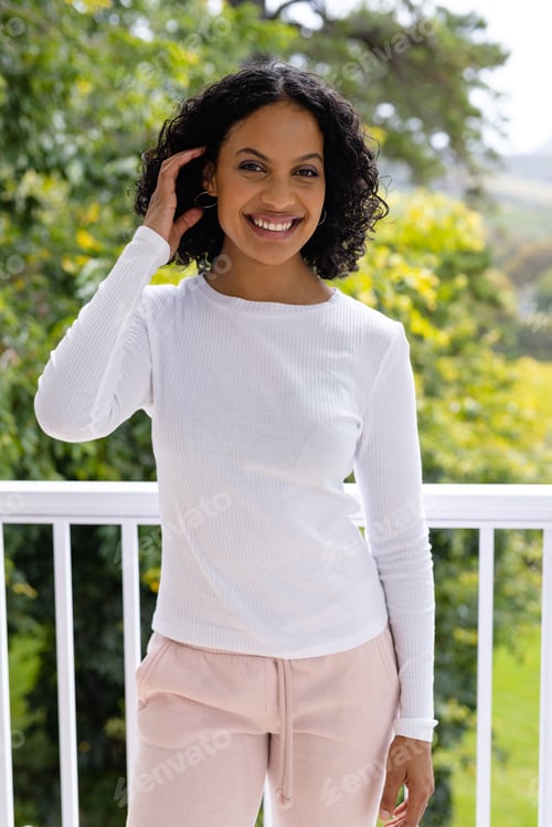 Preview: Portrait of smiling biracial woman standing on sunny balcony at home in front of treetops