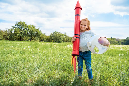 Preview: smiling red hair kid with rocket and astronaut helmet standing in summer field