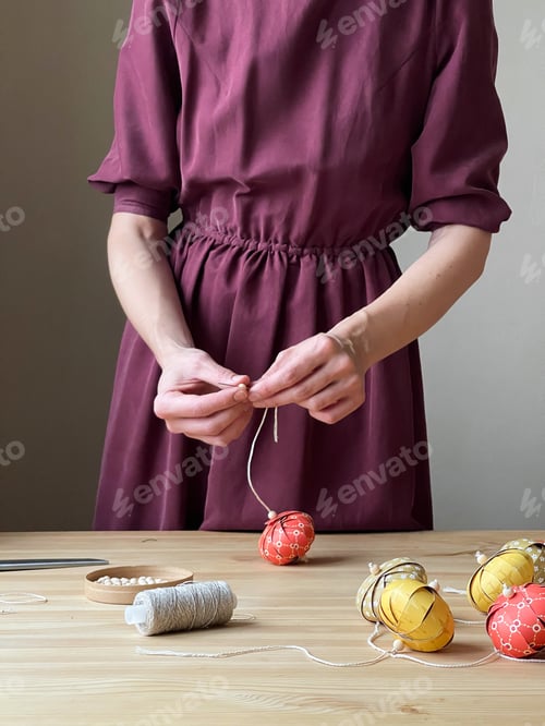 Preview: An unknown woman in a burgundy dress stands at a wooden table and makes paper Christmas balls.