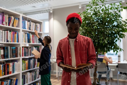 Preview: Smiling student black guy stands in library read book in background girl reach for top of bookshelf.
