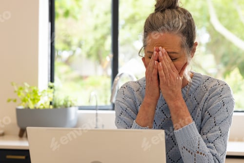 Preview: Stressed mature woman at home covering face with hands in front of laptop