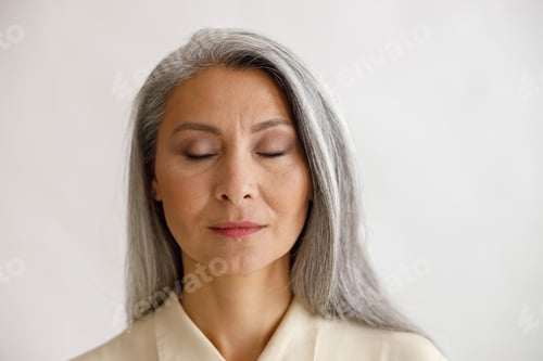 Preview: Tranquil grey haired lady with closed eyes and elegant makeup poses on light background
