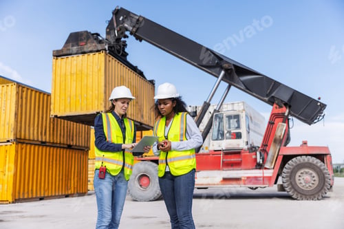 Preview: Two female workers in safety vests and hard hats discuss logistics using a tablet
