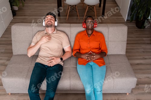 Preview: Two friends relaxing on a couch wearing headphones and enjoying music at home during the afternoon