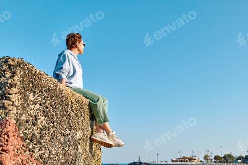 Preview: Young woman sitting on big stone in seaside with blue sky in background and enjoying freedom.