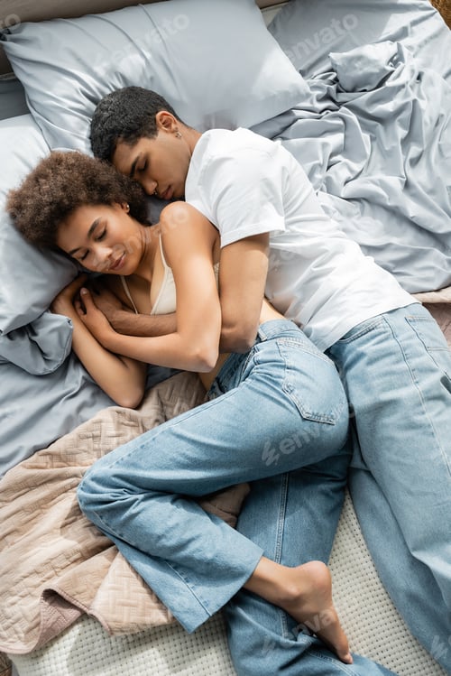 Preview: top view of young african american couple in jeans embracing while sleeping on bed at home