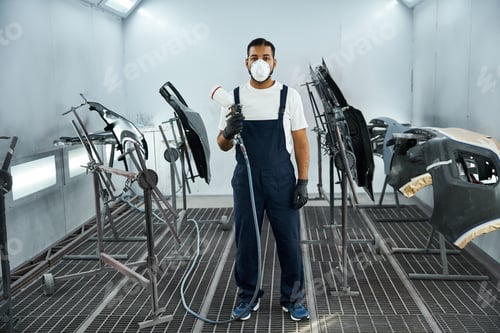 Preview: Handsome young mechanic spraying paint in a well lit workshop with car parts around