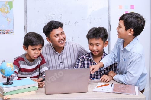Preview: Happy diverse junior school children students gathered at teacher table looking at laptop