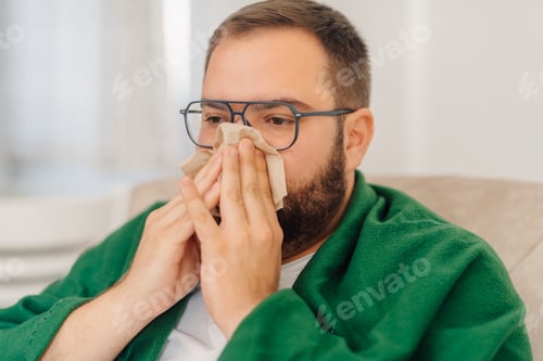 Preview: Sick man blowing nose while sitting on sofa at home