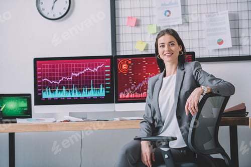 Preview: Smiling computer systems analyst looking at camera near charts on computer monitors on table