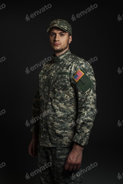 Preview: patriotic American soldier in uniform and cap looking at camera isolated on black